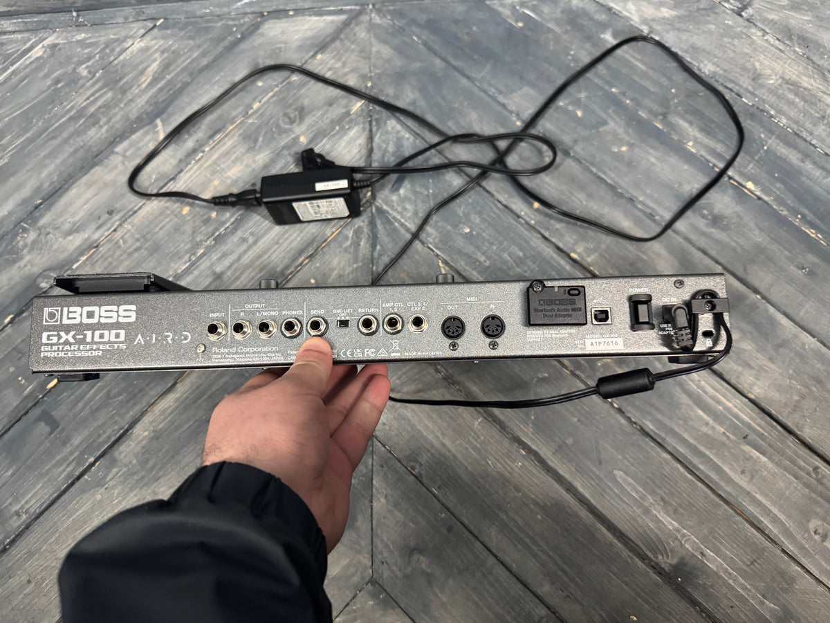 Boss GX-100 guitar effects processor on a wooden surface with a hand holding it.