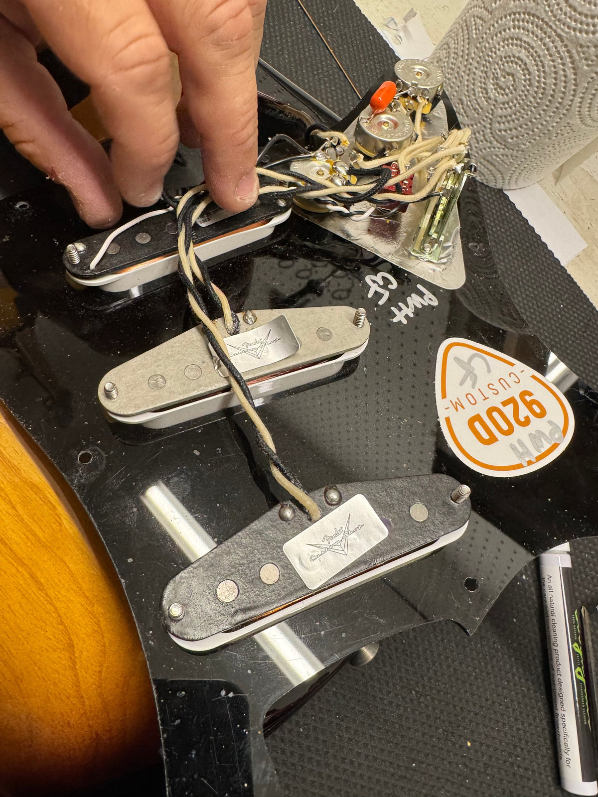 Close-up of guitar pickups and wiring with a hand pointing at them, on a black surface.