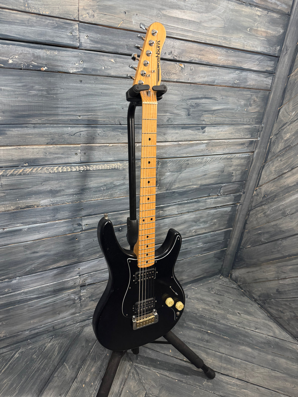 Black electric guitar on a stand against a wooden wall.