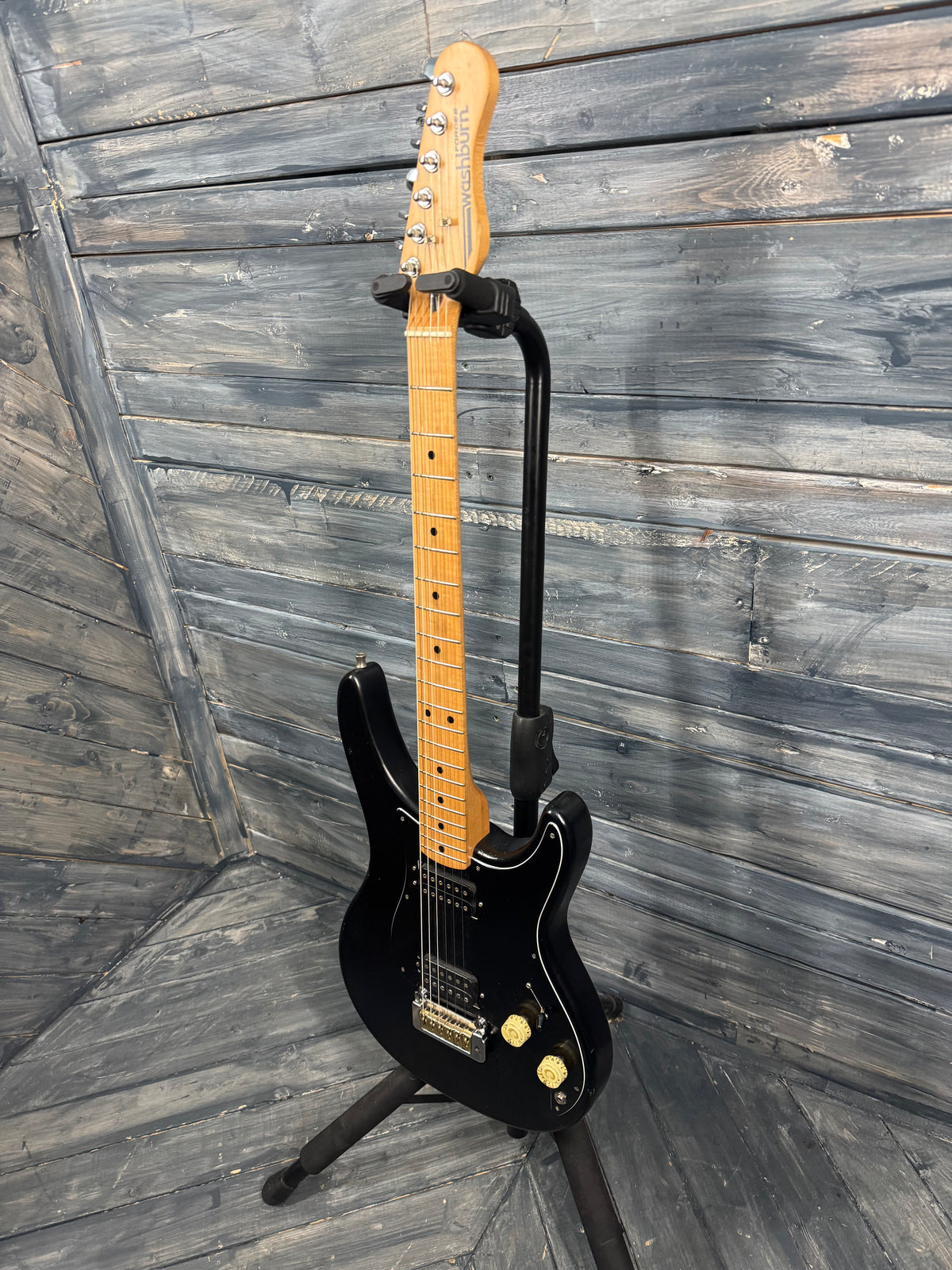 Black electric guitar on a stand against a wooden wall.